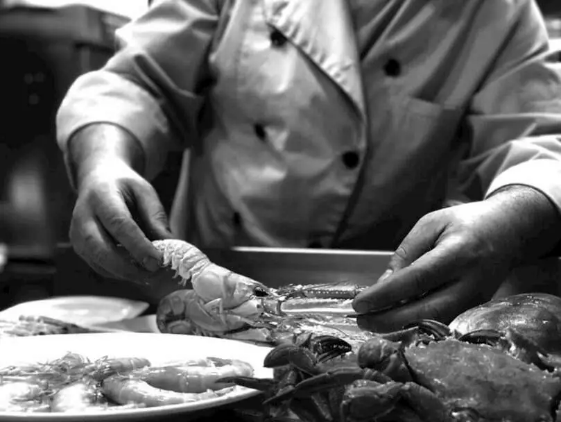 Black-and-white photo of hands in chef whites preparing fresh seafood, arranging langoustine and prawns beside a crab on a plate