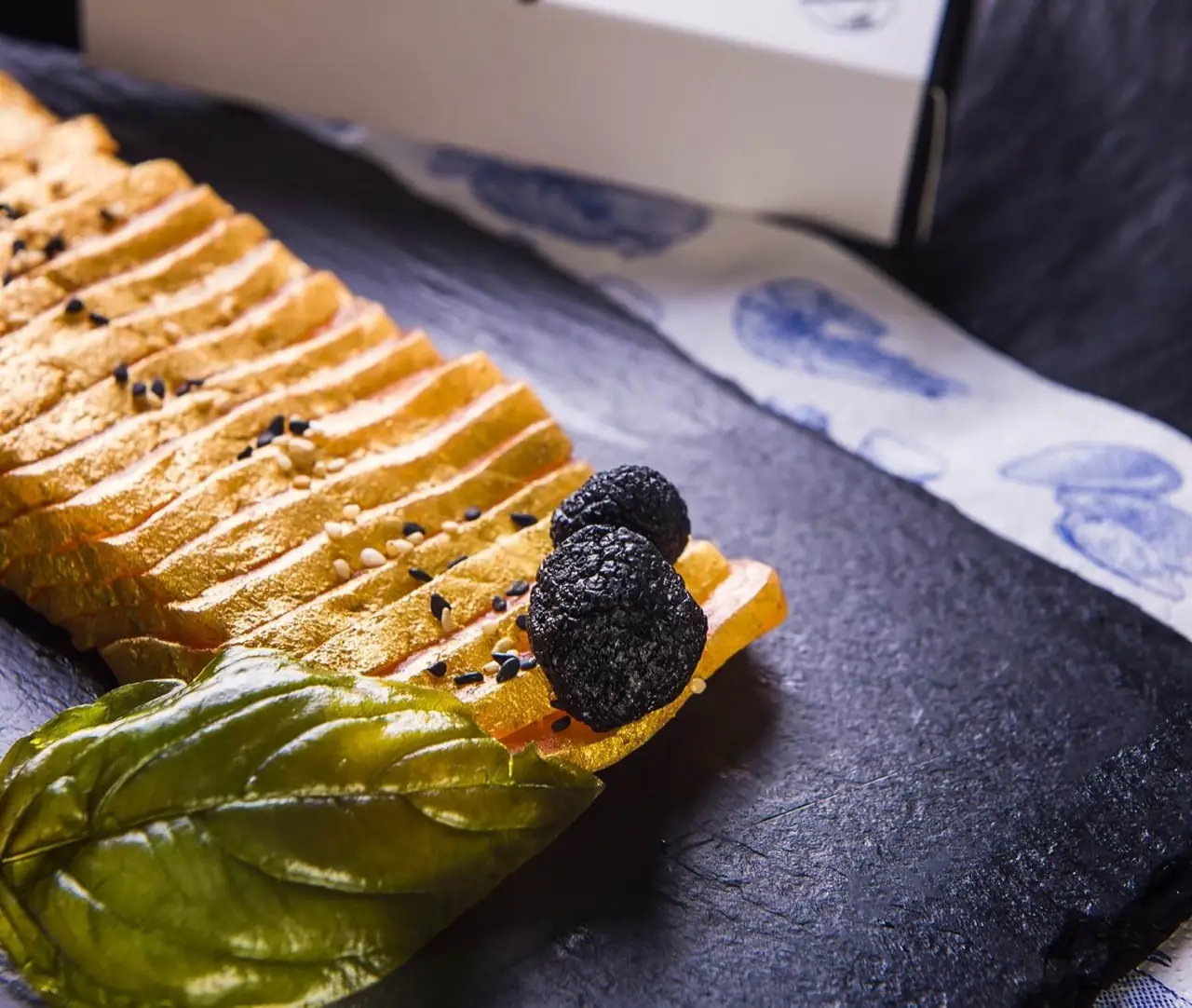 Fanned slices of cured salmon topped with black truffle rounds, sesame seeds and a basil leaf on a slate board