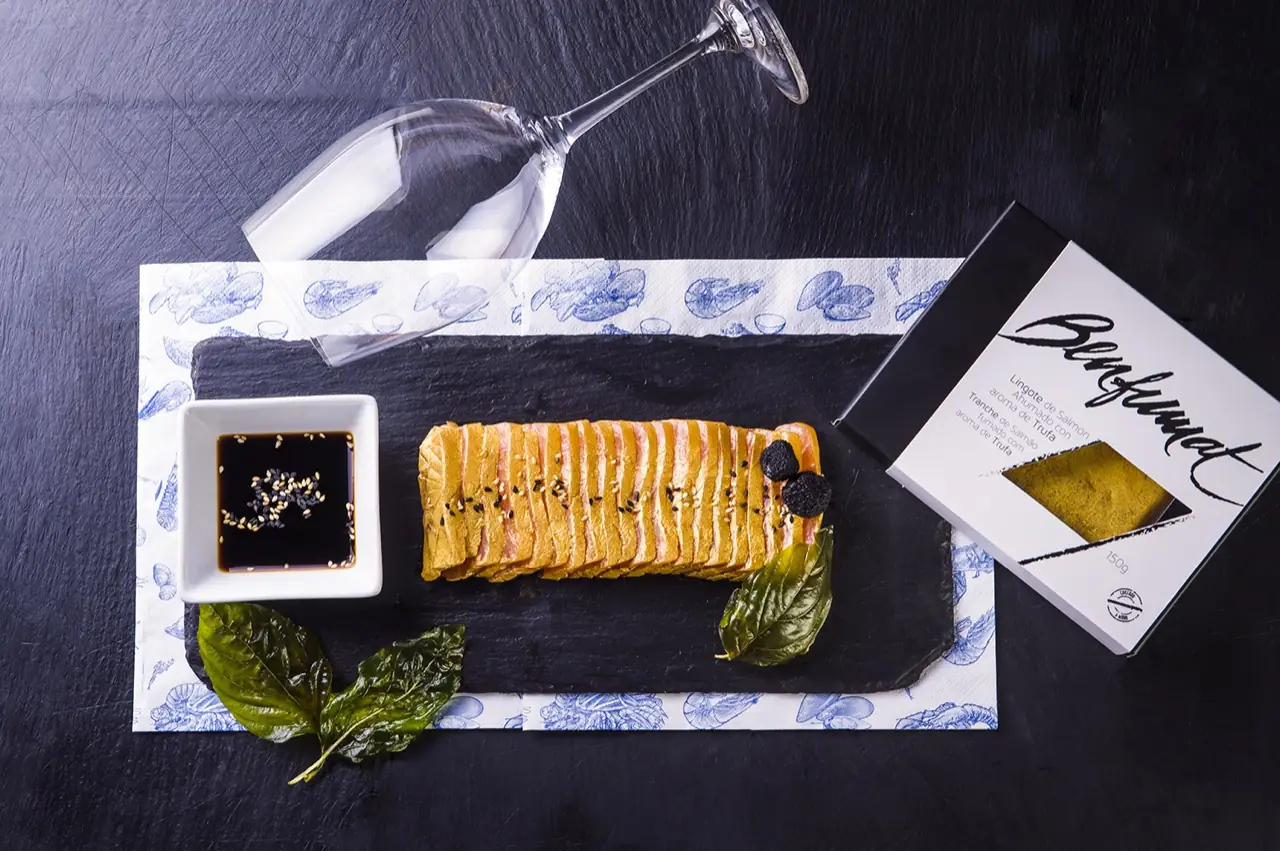 Overhead of sliced smoked salmon loaf with black truffle on slate, a dish of soy with sesame, basil and a wine glass