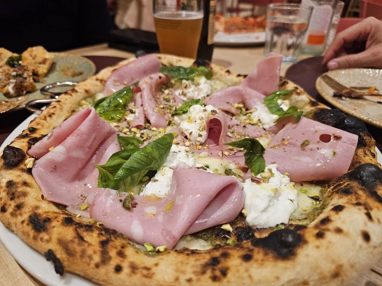 A pizza topped with slices of mortadella and white cream on a table with a glass of beer behind it