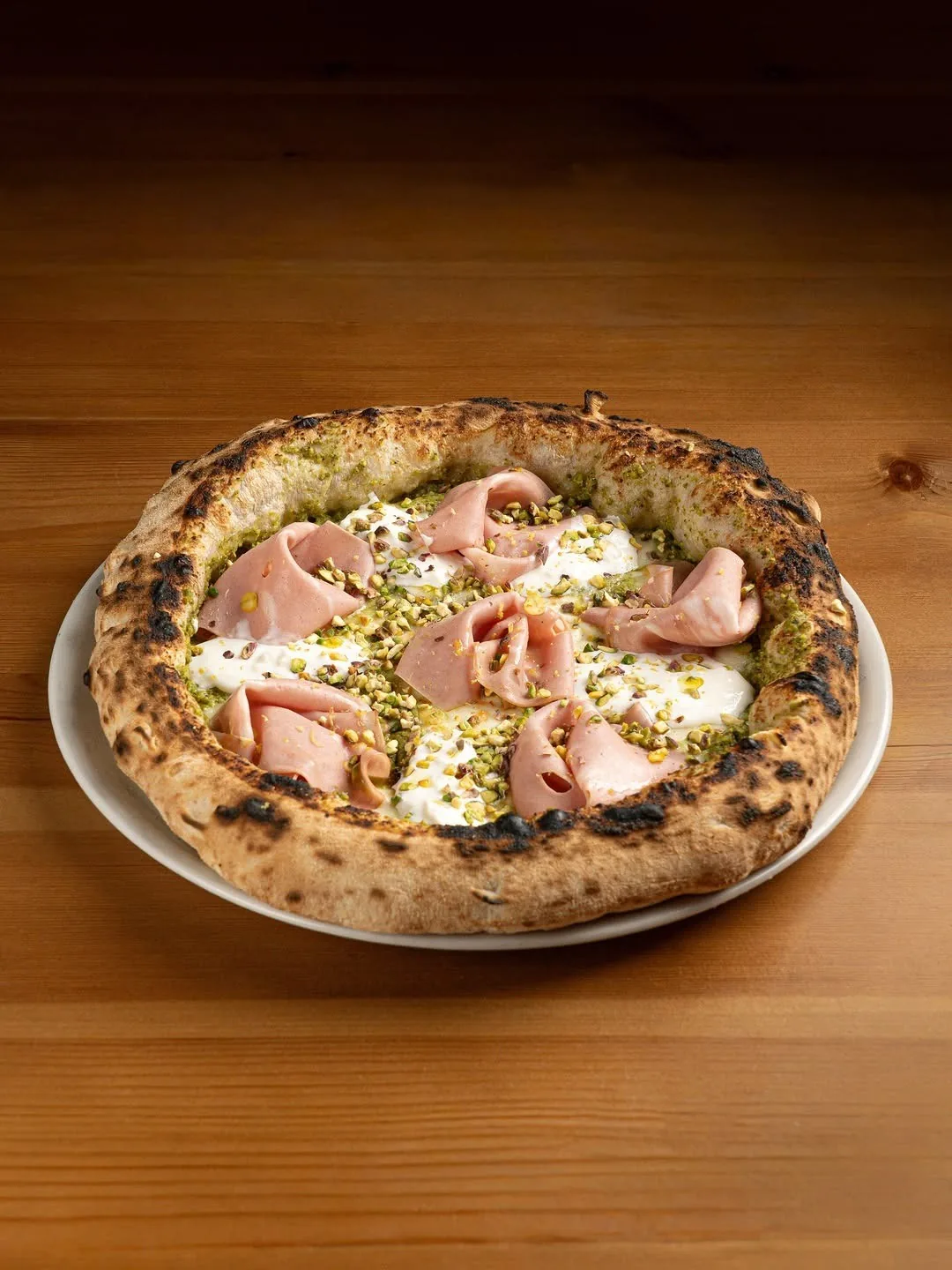 A pizza with slices of mortadella and pistachios on a white plate on a wooden table