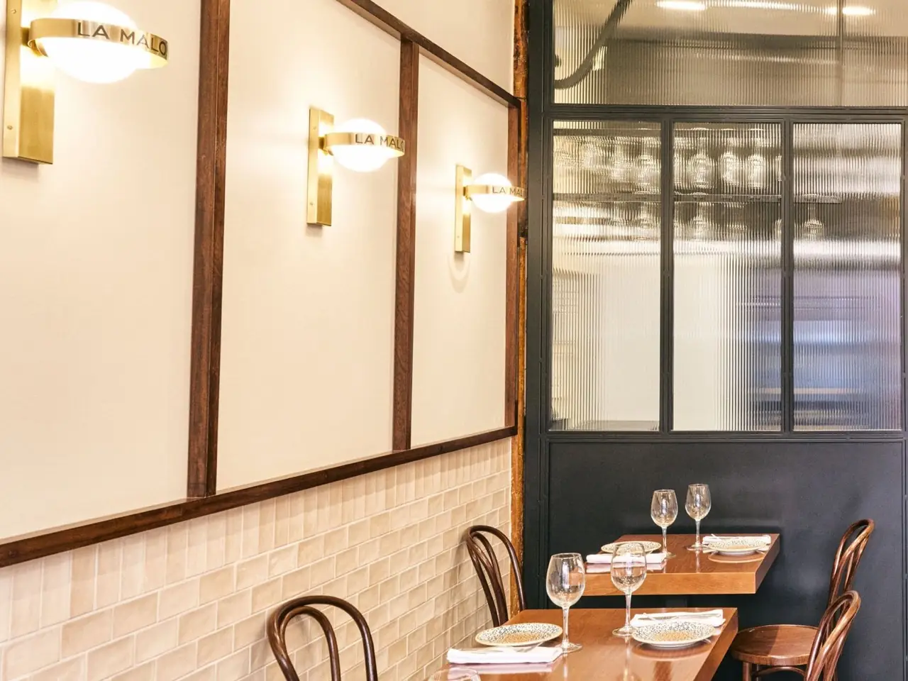 Dining room corner with cream walls, brass wall sconces branded LA MALO, beige subway tile, a reeded-glass partition and set wooden tables with wine glasses and bentwood chairs