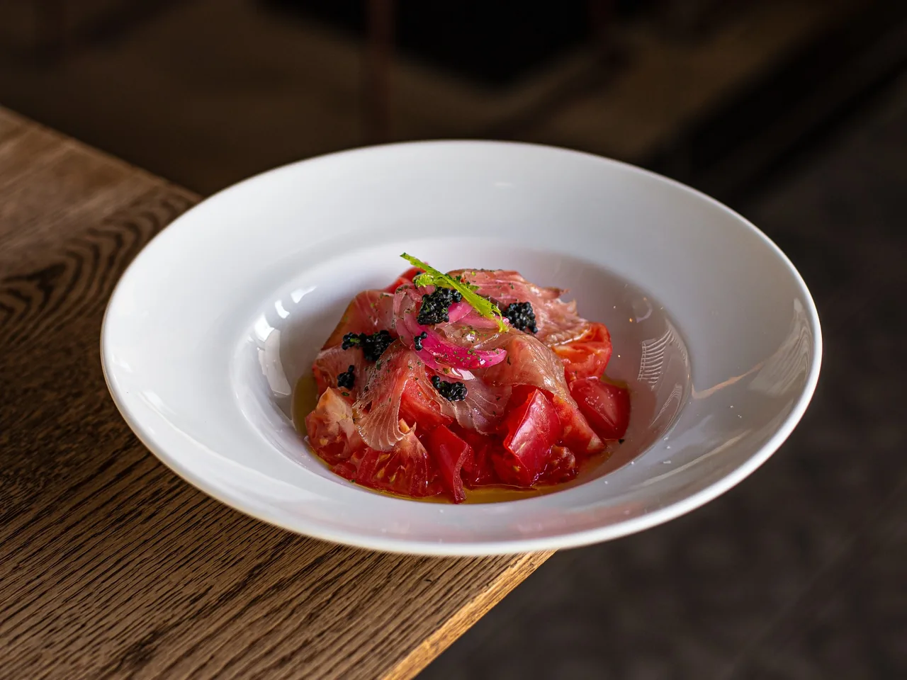 A white bowl of red tomato with thin slices of cured fish and dark pearls on a wooden table