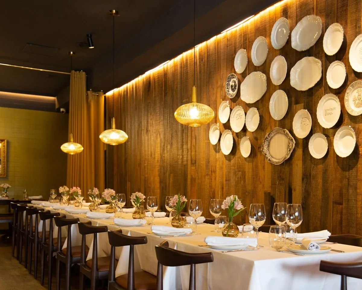 A long set table with white cloths, glasses and flowers beside a wall of decorative plates