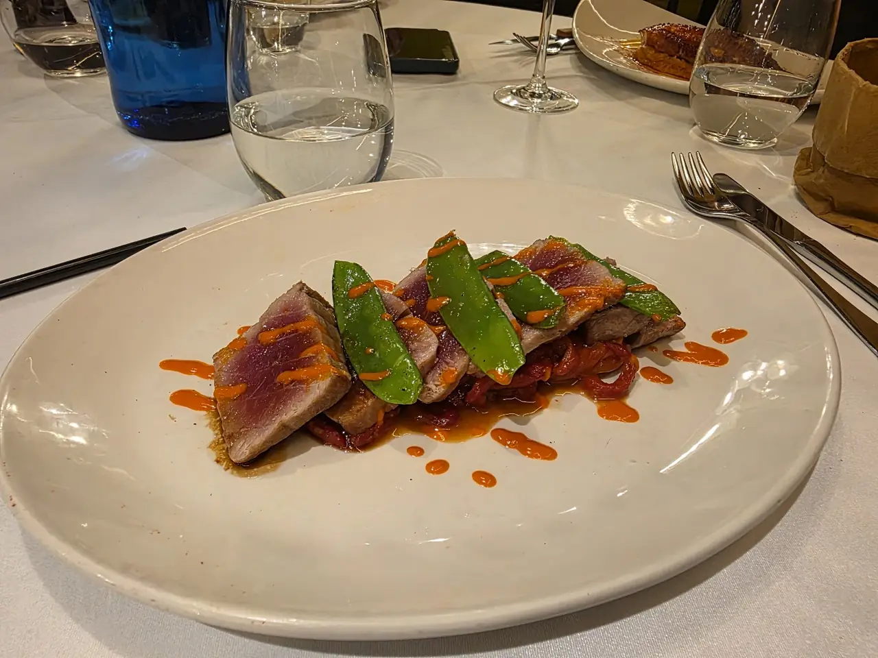 Seared tuna slices with snow peas and roasted pepper strips, drizzled with spicy sauce on a white plate