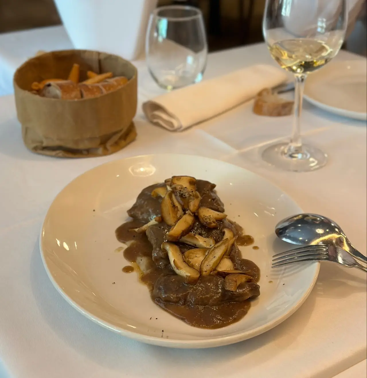 Braised meat in brown sauce topped with sauteed mushrooms on a white plate, with a bread basket in the background