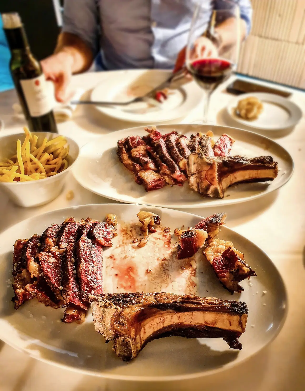 Two plates of sliced grilled T-bone chops with a bowl of shoestring fries and a glass of red wine