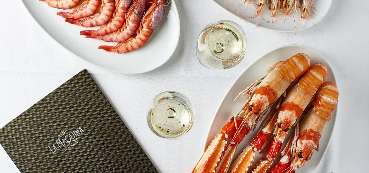 Overhead of a seafood spread on white tablecloth: platters of pink prawns, langoustines and shrimp with two glasses of white wine and a La Maquina menu card