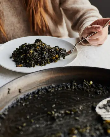 A hand spoons squid-ink black rice (arroz negro) from a paella pan onto a white plate