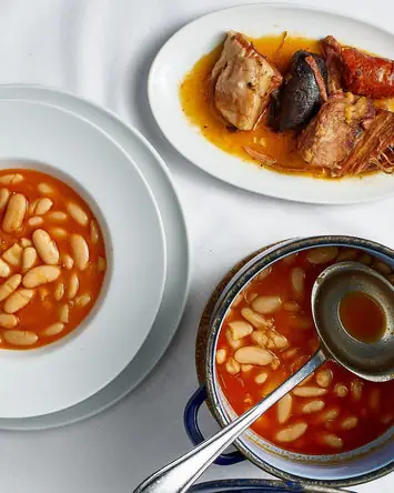 Fabada: a bowl of white beans in broth, a plate of stewed meats and chorizo, and a blue pot with ladle