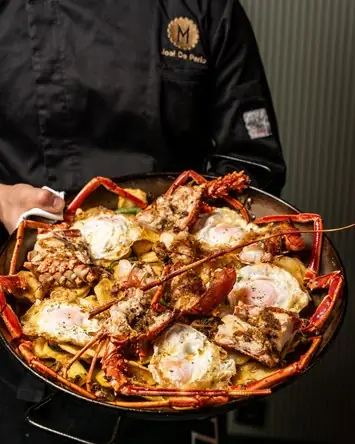 A large pan of fried eggs with lobster and langoustine over potato crisps, held by staff in a black jacket
