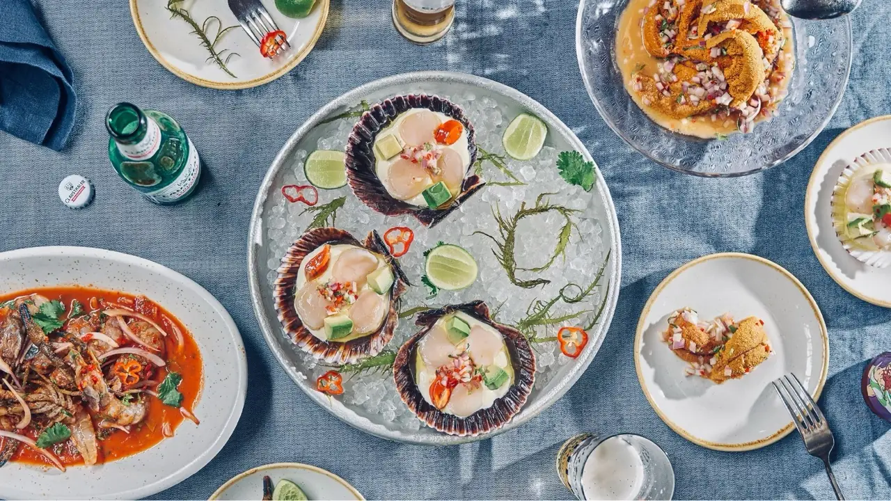 Wide overhead shot of a denim-covered table set with scallop-shell ceviche on ice, a bowl of tiger's milk seafood stew, a plate of sea urchin tiradito, and glasses of beer.