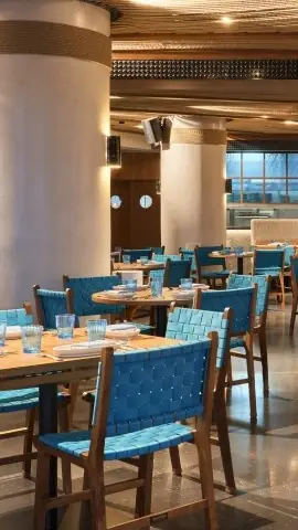 Restaurant dining room with woven blue chairs, round wooden tables set with glasses, and stone columns under warm lighting.