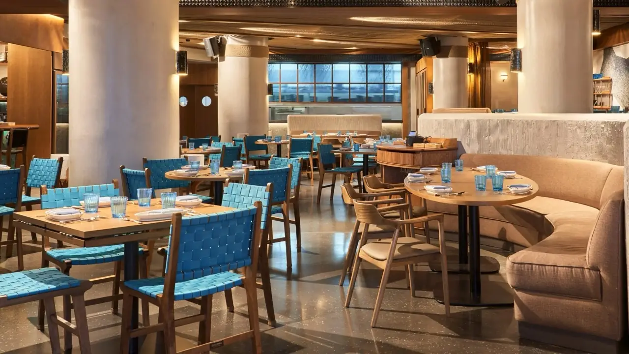 Wide view of a restaurant dining room with woven blue chairs, round tables, a curved banquette, stone columns, and an open kitchen visible through a window in the back.