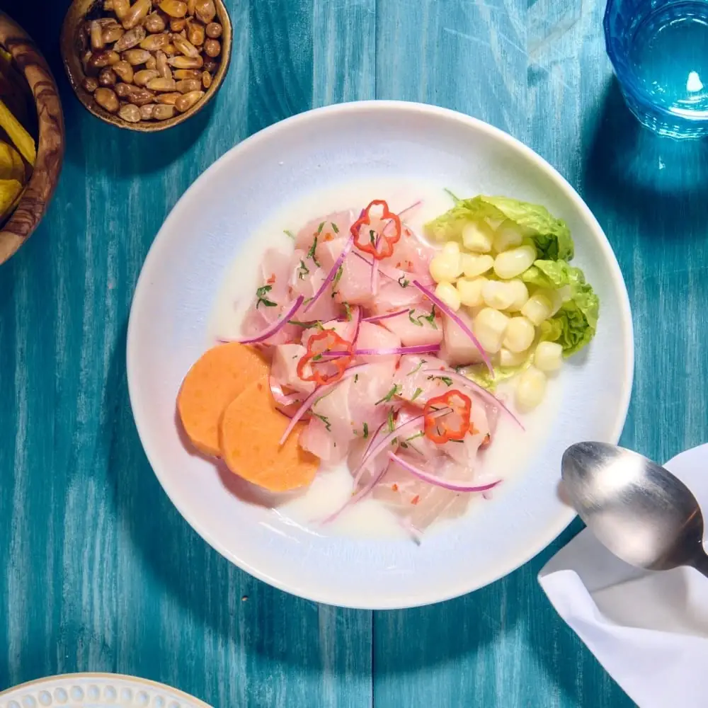 Overhead shot of a ceviche plate with white fish, red onion, corn, lettuce, sweet potato slices and chili on a blue wooden table, with a small bowl of toasted corn nuts alongside.