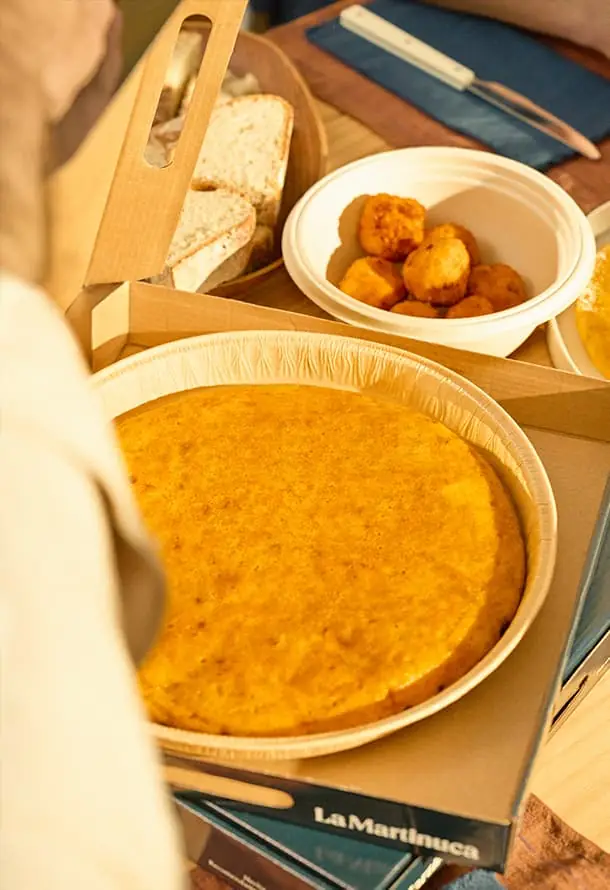 Whole Spanish potato tortilla in a wooden takeaway tray beside a bowl of golden croquettes and sliced bread, on a branded La Martinuca box
