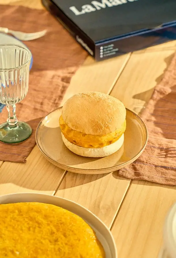 Tortilla sandwiched in a soft white bread bun on a beige plate, with a ribbed water glass and a La Martinuca box on a wooden table