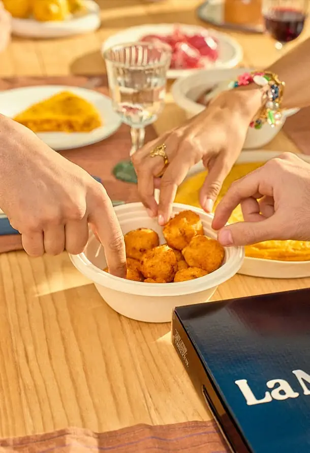 Three hands reaching into a bowl of golden fried croquettes on a wooden table set with tortilla, wine and small plates
