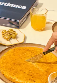 Whole golden tortilla being sliced with a knife, beside a skewer of olives and a glass of cider, with a La Martinuca box behind