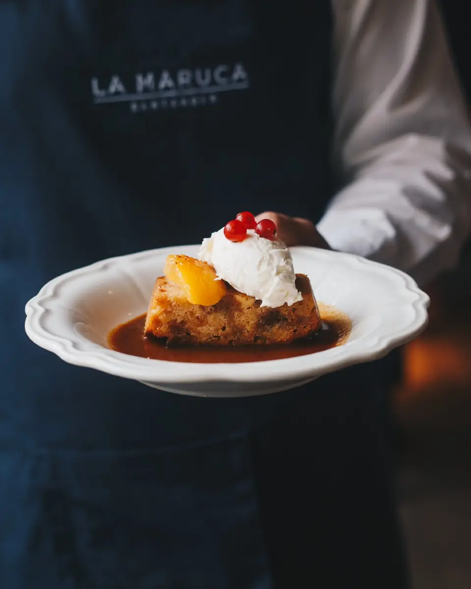 Sticky toffee cake with whipped cream and red currants on a white plate, held by a server