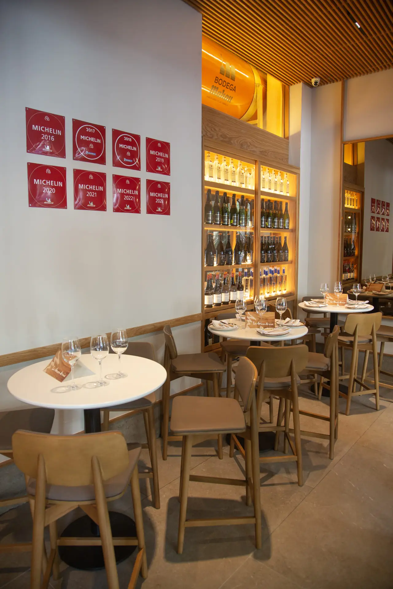 The bar room with round white tables set with wine glasses and La Monteria table cards, a lit wooden wine shelf behind and eight red Michelin plaques dated 2016 to 2023 on the side wall
