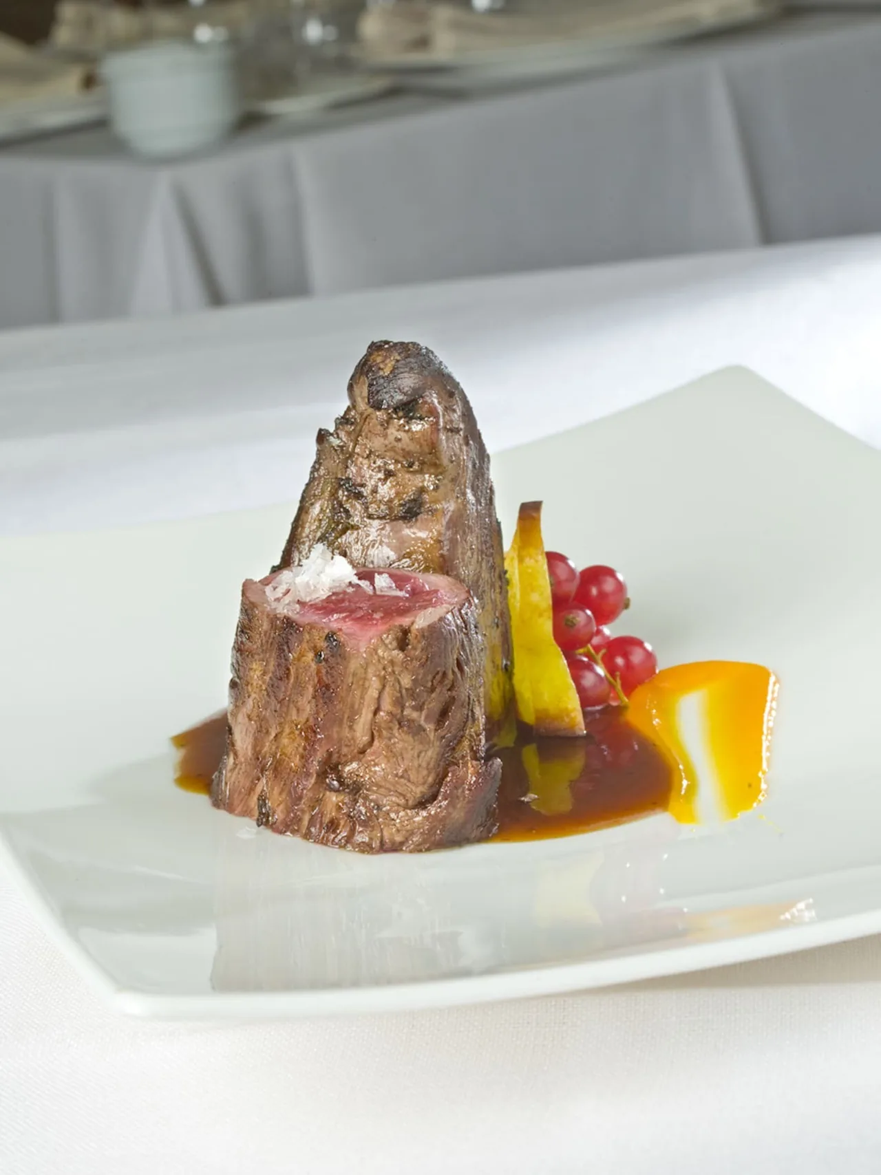 A tall piece of roasted meat with red currants and small vegetables on a white plate