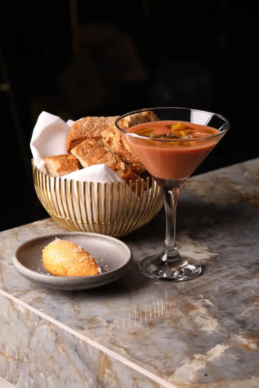 Chilled tomato soup topped with diced garnish served in a martini glass, with a gold bread basket and a butter quenelle on marble