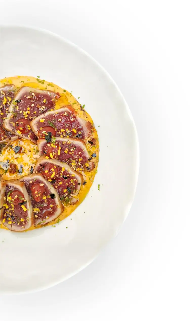Slices of seared red tuna fanned over an orange sauce with sesame and herbs on a white plate