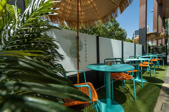 Outdoor terrace with teal round tables, orange woven chairs and straw parasols on artificial turf, La Morena logo on the wall