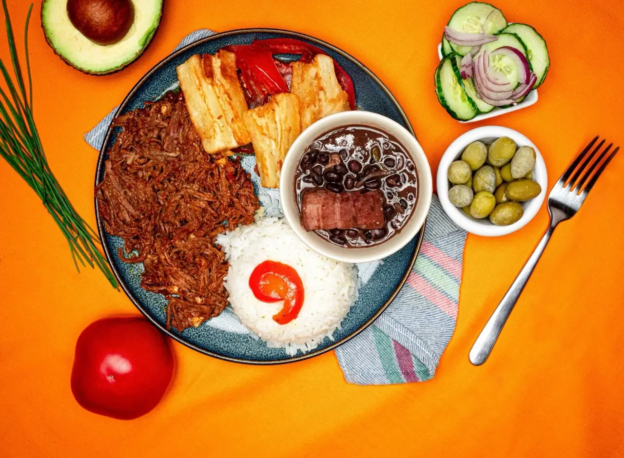 Overhead of a Latin American plate with shredded beef, white rice, fried yuca and a bowl of black beans with a sausage, plus side dishes of olives and cucumber-onion salad on an orange cloth