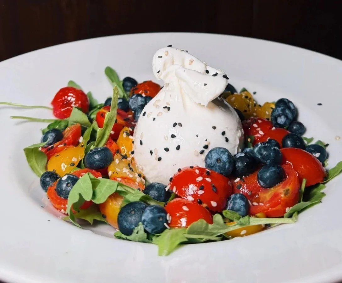A white plate of burrata with fresh tomatoes and blueberries