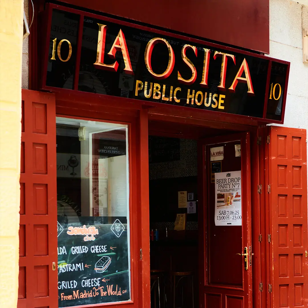 Red-framed storefront of La Osita Public House with an illuminated red-and-gold sign and a chalkboard listing grilled cheese and pastrami sandwiches