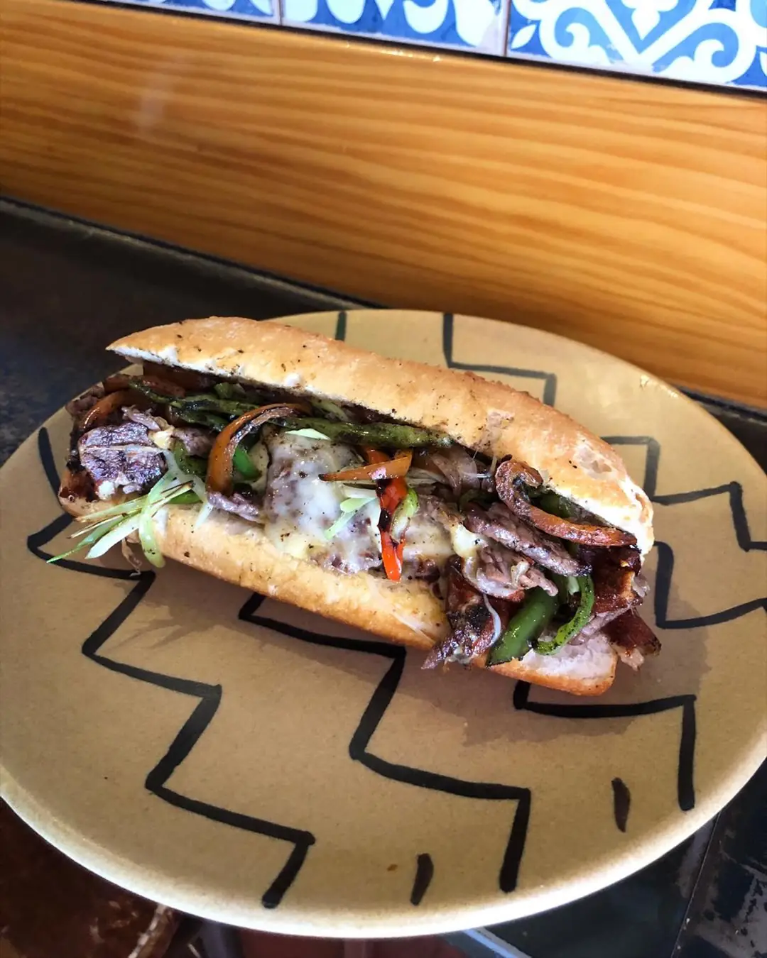 A grilled steak sandwich with melted cheese, peppers, and greens on a patterned plate