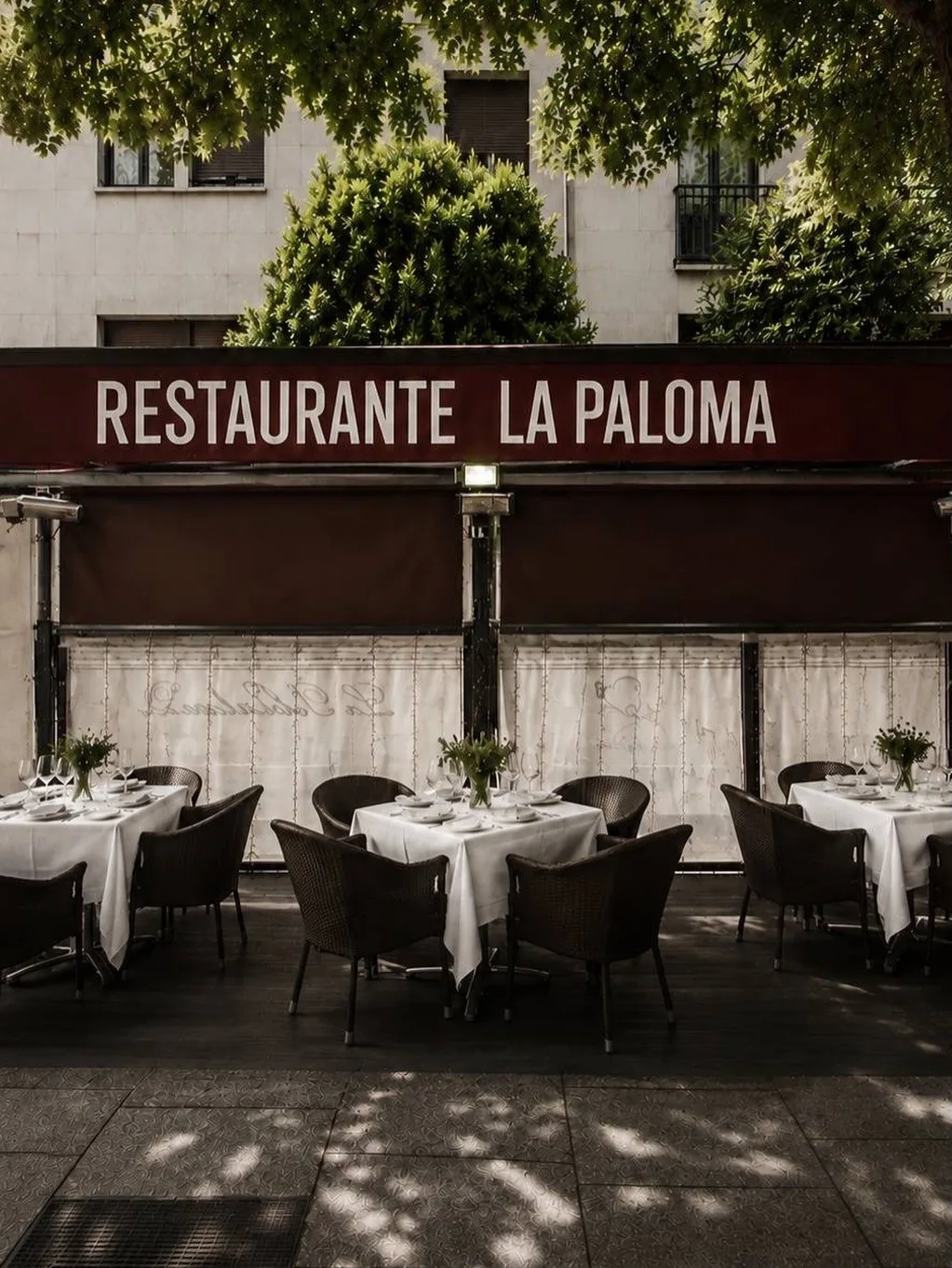 Tables laid with white tablecloths on an outdoor terrace below a red awning with the restaurant name
