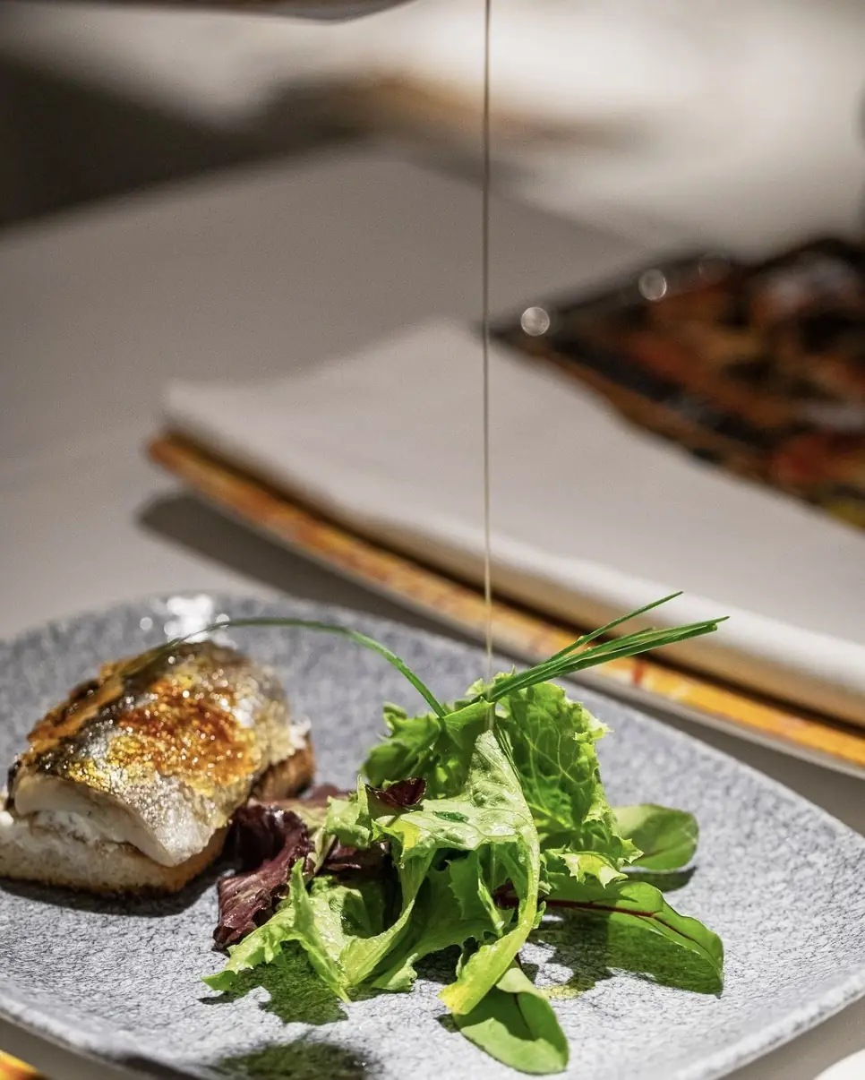 Seared fish fillet with mixed greens on a textured plate, olive oil being drizzled over it