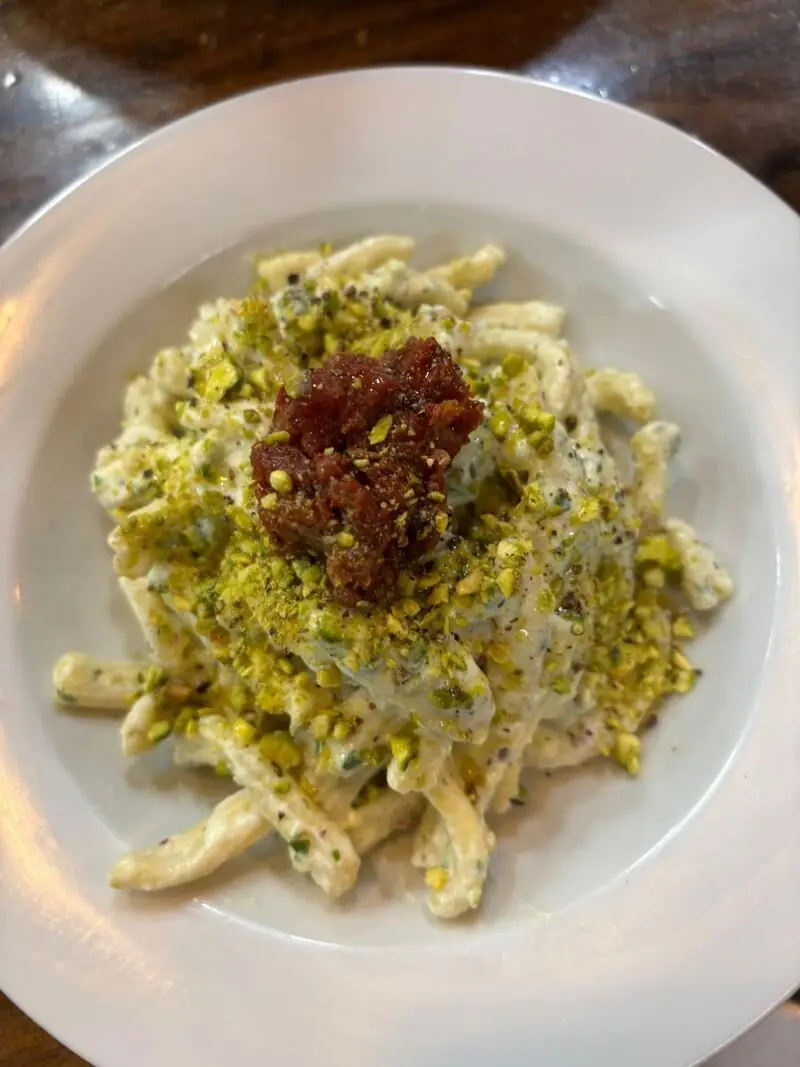 White bowl of creamy pasta dusted with crushed pistachio and topped with a spoonful of red jam or cured meat