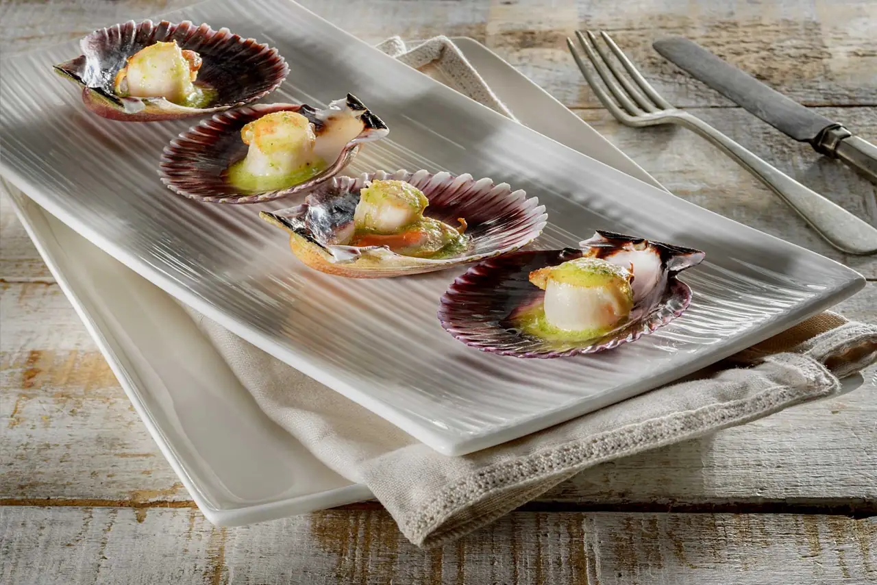 Four grilled scallops in their shells topped with green sauce, arranged in a row on a long ribbed white platter with a fork and knife beside it.