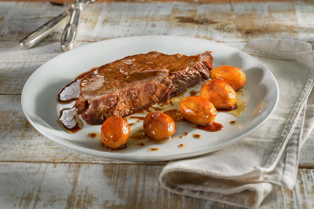 Sliced braised beef in a dark sauce with glazed baby potatoes on a white plate.