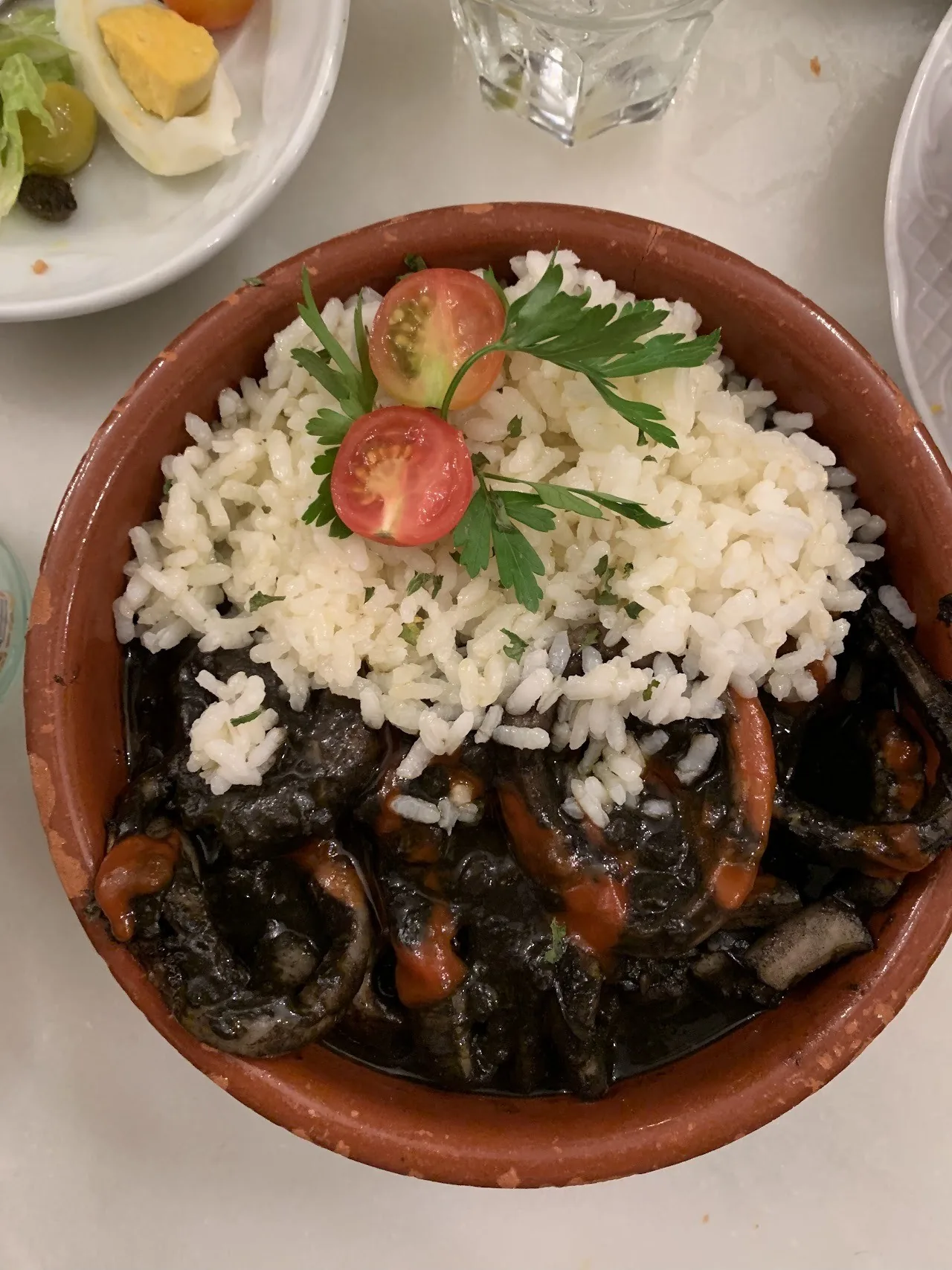 A terracotta dish of white rice with dark seafood and sliced tomato