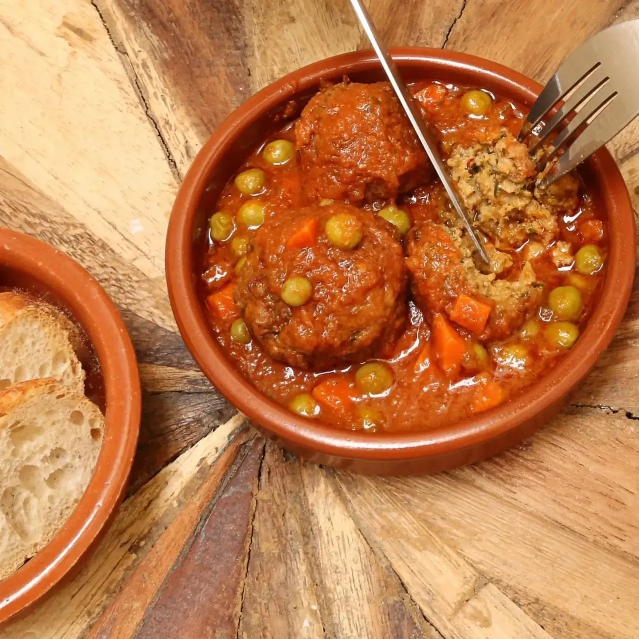 Vegan meatballs in tomato sauce with peas and carrots in a terracotta dish, with bread on the side, at La Perra Verde