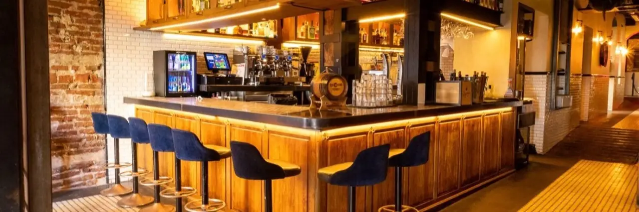 Interior of a bar with a curved wooden counter, blue velvet stools, exposed brick wall, coffee machine and vermut barrel behind the bar