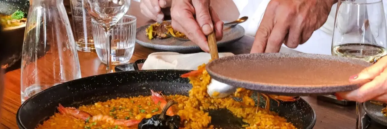 Black paella pan of saffron rice with whole prawns and a black chilli, being served onto a stoneware plate at a table with wine glasses