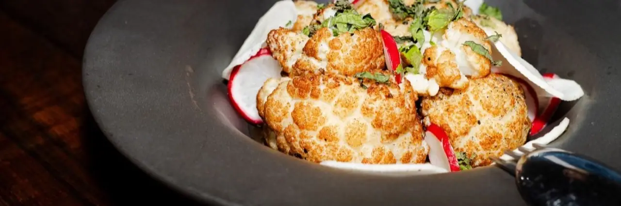 Roasted golden cauliflower florets with sliced radish and chopped herbs in a dark stoneware bowl