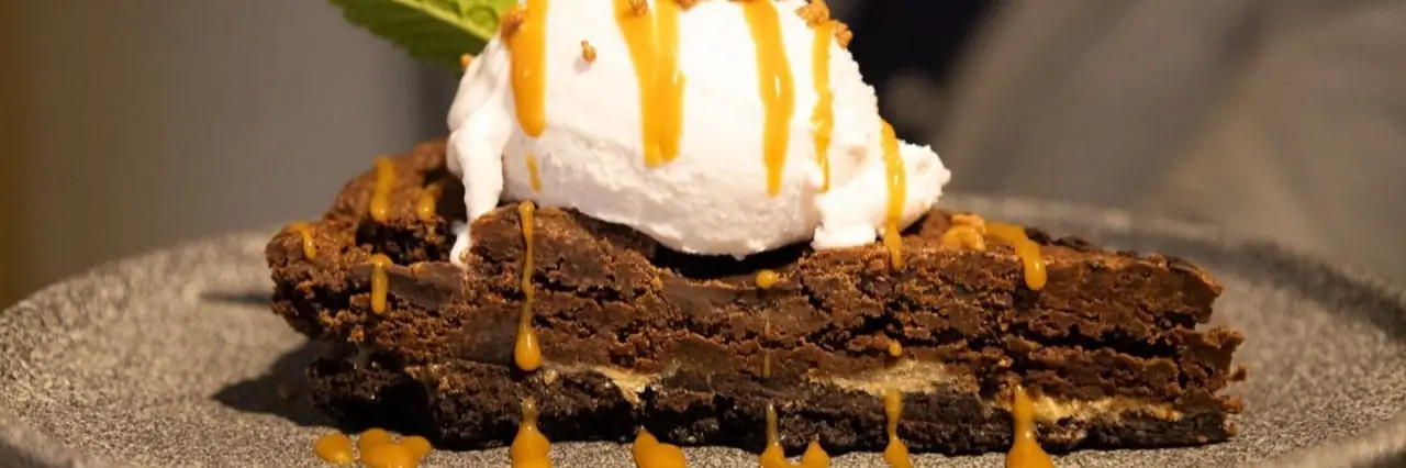 Chocolate brownie slice topped with a scoop of ice cream and drizzled with caramel sauce on a grey plate