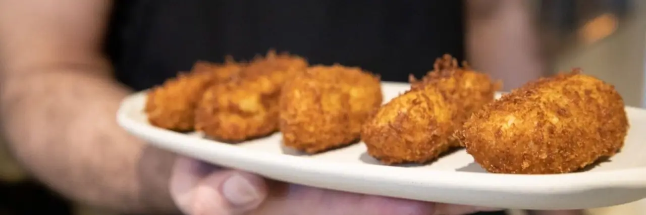 Five golden fried croquetas lined up on a white plate held by a person