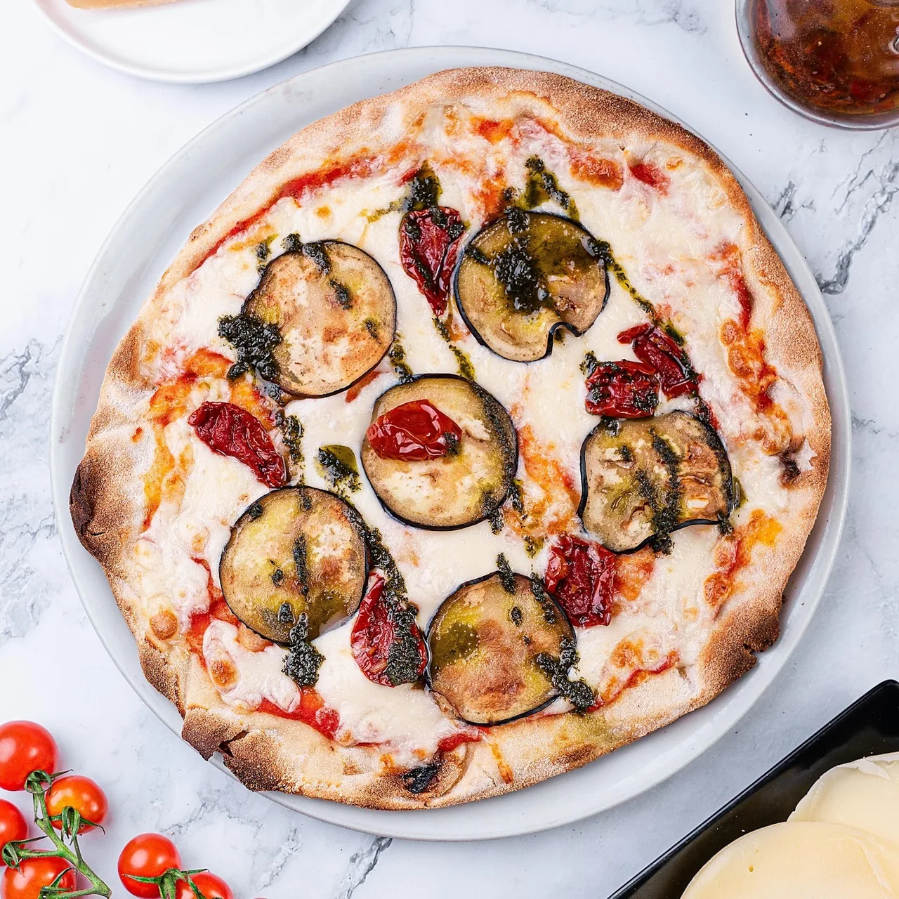 A pizza topped with grilled aubergine slices, sun-dried tomatoes and pesto on a white plate on a marble table
