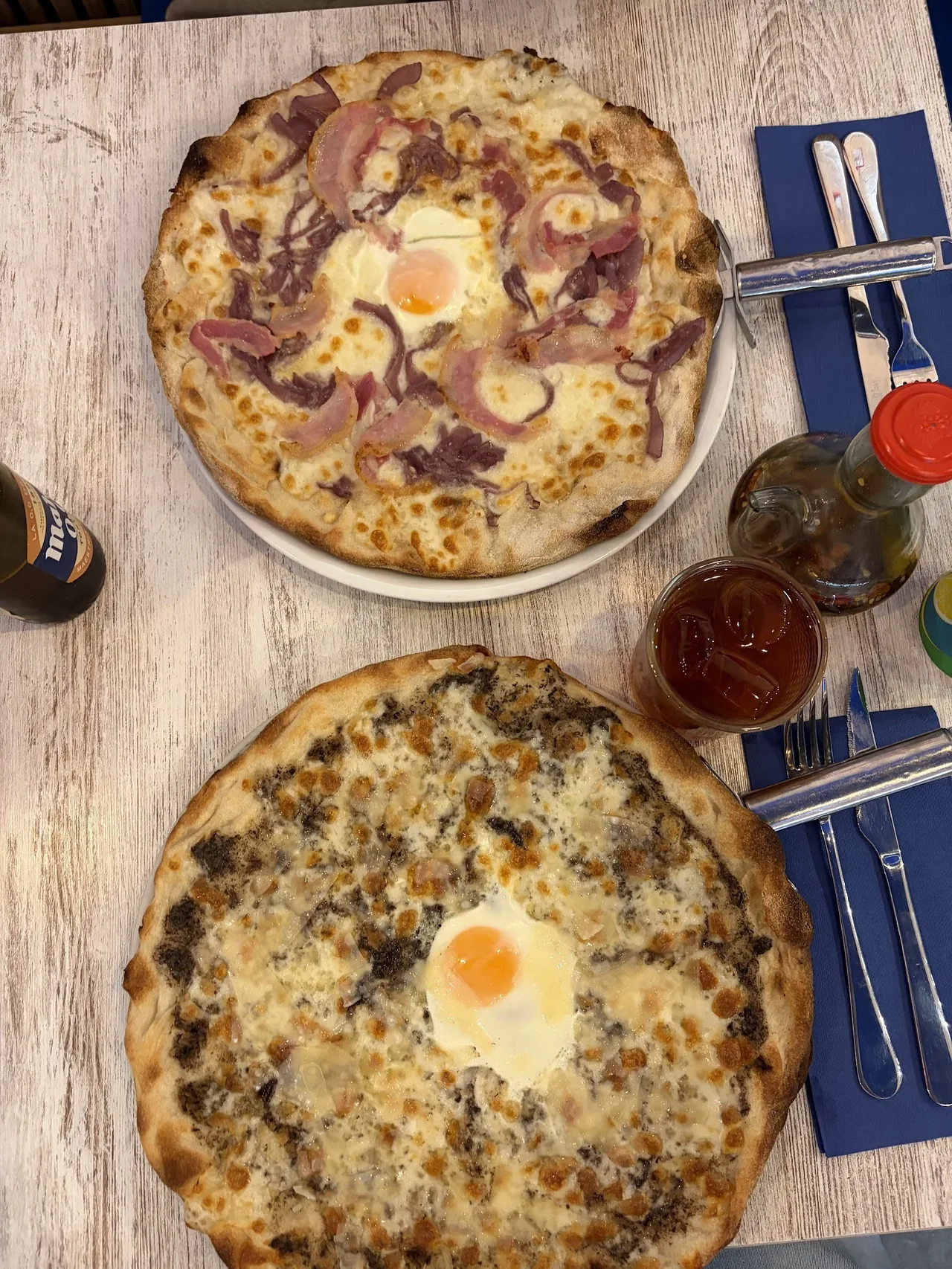 Two pizzas seen from above on a light wooden table, one with an egg in the centre