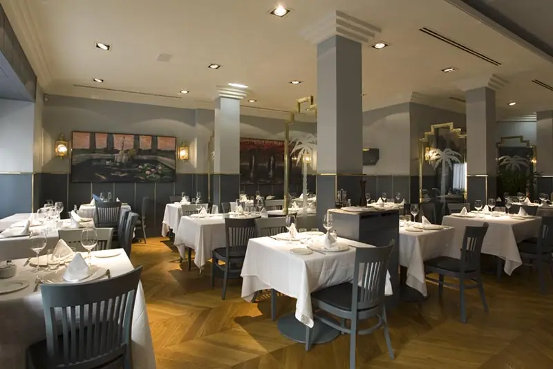 Elegant restaurant dining room with white tablecloths, grey upholstered chairs, herringbone wood floor, and framed artwork on grey walls with brass column trim.