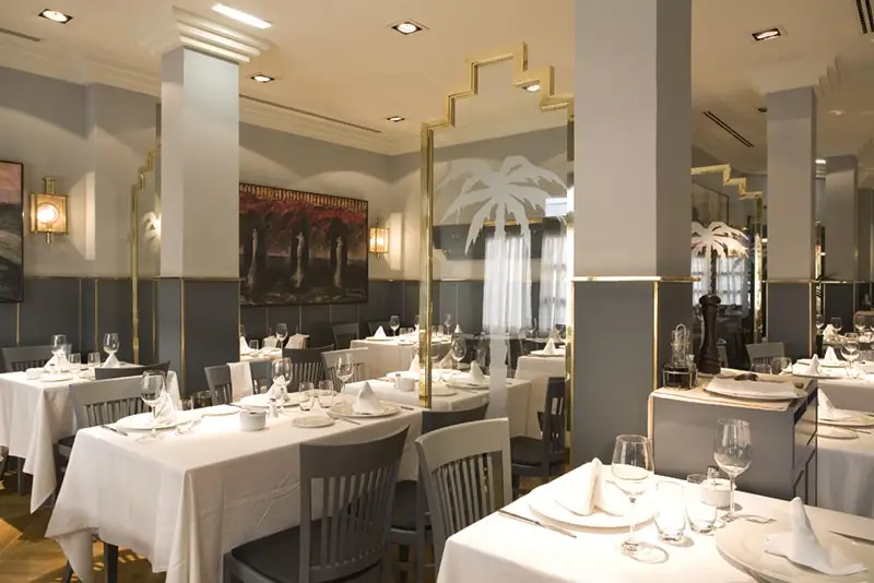 Restaurant dining room with white-clothed tables set with wine glasses, a decorative frosted-glass palm-tree divider, and framed artwork on the wall.