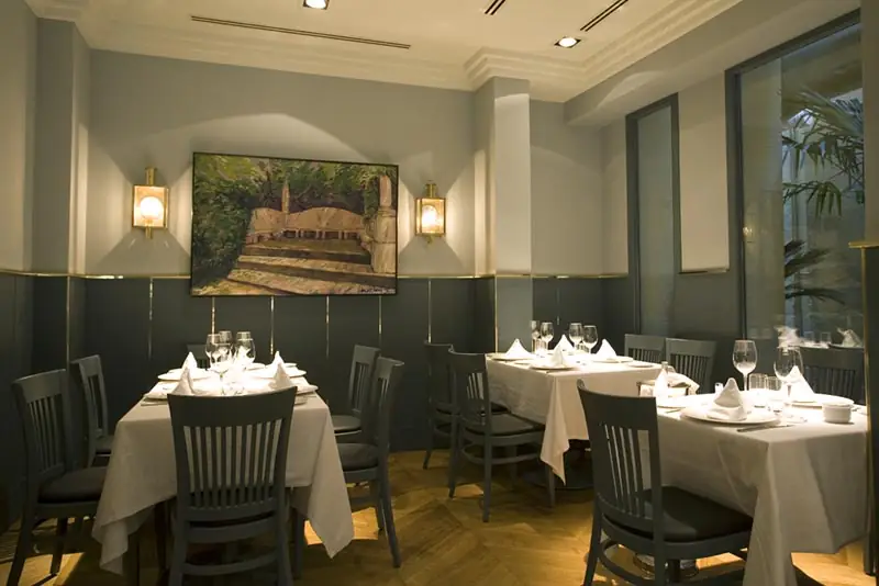 Restaurant dining room with white-clothed tables, grey chairs, wall sconces flanking a landscape painting, and a view into a plant-filled area near the window.